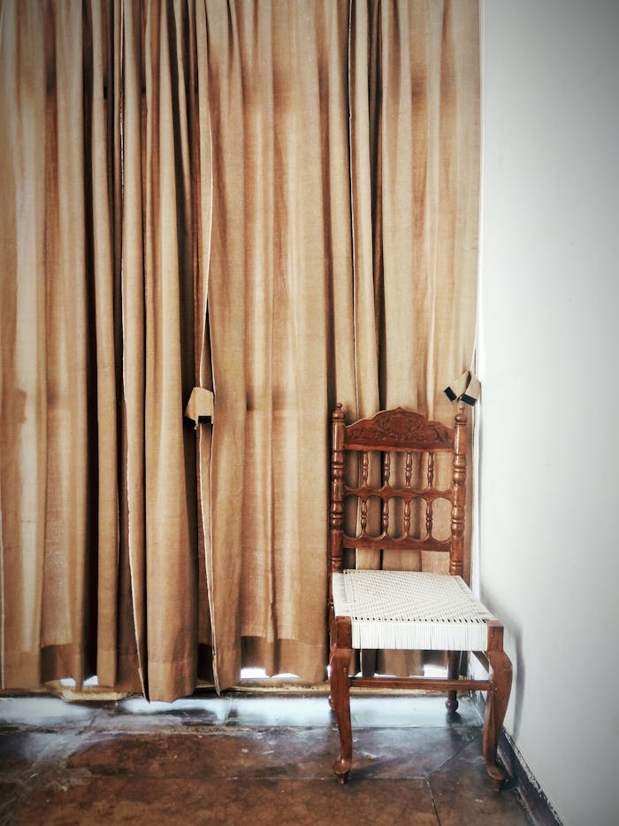 A classic wooden chair next to brown curtains in a sunlit room.